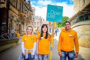 Three students in orange t-shirts with Open Day placard on campus