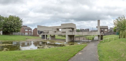 External photo of large abstract building consisting of concrete blocks , set across a pond in a setting of open grass area and surrounding houses