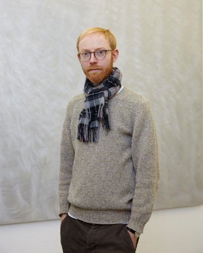Mid-shot photo of male, slim, red hair, grey sweater and wearing blue tartan scarf against grey background