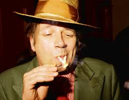Close-up colour portrait photo of white middle-aged man in a tan fedora hat , cark suit, red shirt and tie, lighting a cigarette with a match