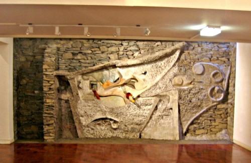 Interior setting of large abstract wall sculpture set on a wall of slate stones, with wooden roof beam across top of picture and wooden flooring in foreground